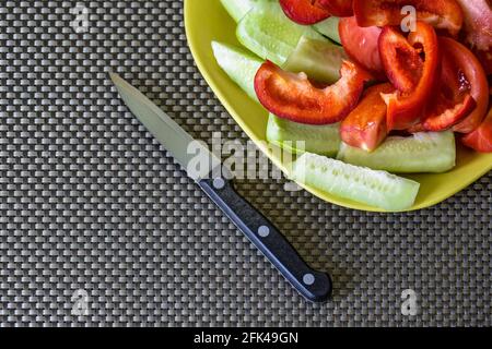 Vista dall'alto di verdure fresche a fette - cetrioli, pomodori e peperoni dolci su piastra gialla in ceramica e coltello da cucina. Sfondo verde scuro. Foto Stock