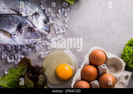 Cornice di ingredienti crudi per cucinare cibi gustosi e sani. Pesce dorado fresco su ghiaccio, verdure, uova su fondo grigio in cemento vista dall'alto. Foto Stock
