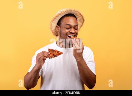 Buon ragazzo afroamericano in abito estivo informale mangiare gustosa pizza italiana, gustando il suo spuntino su sfondo giallo Foto Stock