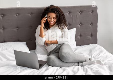 Donna nera felice seduta a letto che parla al telefono Foto Stock