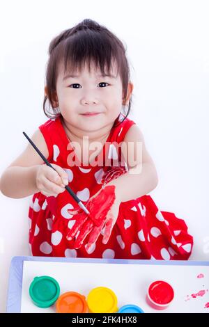 Piccolo bambino asiatico (tailandese) felicemente, bella ragazza che guarda la macchina fotografica, sorridendo e dipingendo la sua mano utilizzando strumenti di disegno multicolore (acqucolo Foto Stock