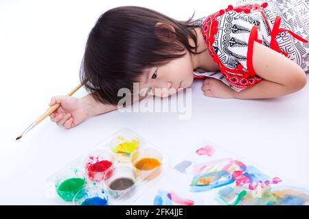 Bambina asiatica (tailandese) sdraiata sul pavimento dello studio e che tiene il pennello. Idea di pensiero bambino per la pittura di opere d'arte, su sfondo bianco. Scatto in studio Foto Stock