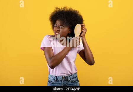 Infastidita donna afroamericana che ha problemi a spazzolare la sua riccia bush capelli su sfondo giallo studio Foto Stock