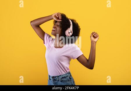 Donna afro-americana spensierata con cuffie wireless che ballano e si diverta con la musica a occhi chiusi, sfondo giallo Foto Stock