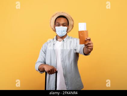 Ragazzo nero millenario con valigia che indossa maschera facciale, mostrando passaporto e biglietti aerei alla fotocamera, sfondo arancione Foto Stock