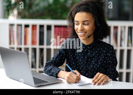 Soddisfatto amichevole giovane afroamericano intelligente con le cuffie, lavoratore di ufficio o operatore di centro di supporto, siede alla sua scrivania, conduce la consultazione in linea con il cliente, prendendo le note, sorrisi Foto Stock