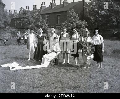 Anni '50, storico, su un pezzo di terra alla fine della terrazza di case vittoriane, un gruppo di uomini locali - 'The Heavy Gang' vestito da donna e vestito fantasia in piedi per una foto prima di uno spettacolo per il festival 'May Day', Inghilterra, Regno Unito. Gli attori dilettanti qui sono in costumi diversi, tra cui Guinevere, la leggendaria Regina di Re Artù. Il giorno di maggio era un'antica tradizione che celebrava l'arrivo della primavera e dell'estate e gli artisti si vestirebbero e rievocavano il folklore antico e le storie danzando intorno ad un palo di legno alto, un maypole, un'attività popolare. Foto Stock