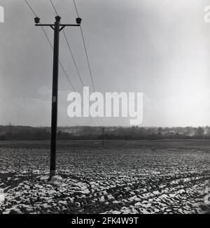 Anni '60, scena storica e invernale, che mostra un palo telegrafico in legno nel mezzo di neve gelido, agitato agricolo o terreno agricolo, con linee telefoniche o fili che lo attraversano ad un'altezza elavata, Inghilterra, Regno Unito. Molti paesi hanno messo le linee telefoniche sottoterra, ma il Regno Unito è uno di coloro che le hanno ancora sopra terra. Credete o no, ci è una società di apprezzamento per i poli del telegrafo. Foto Stock