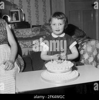 anni '60, storico, su un divano in una stanza anteriore, una bambina seduta con sulla sua torta di compleanno, con quattro candele accese e un piccolo elefante sulla parte superiore. Sembra più come una torta di nozze, ma forse questo era lo stile delle torte di compleanno dei bambini in questa era, Foto Stock