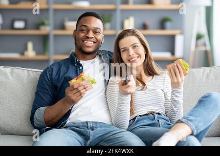Happy giovane coppia multiculturale guardare la TV e mangiare panini a. Casa Foto Stock