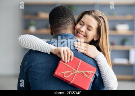 Regalo romantico. Ragazza grata abbracciando il suo ragazzo nero e Holding presente Box Foto Stock