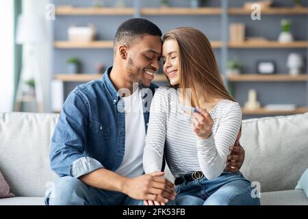 Diventare genitori. Coniugi romantici multirazziali con test di gravidanza positivo a casa Foto Stock