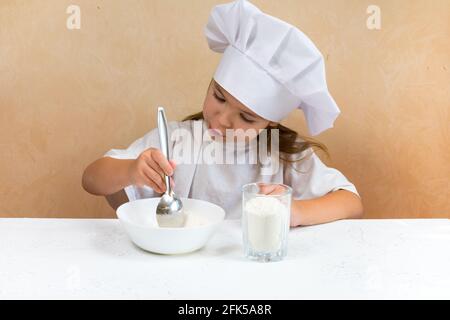 Una bambina vestita come cuoco impasta l'impasto. Cucina bambino stile di vita concetto. Il capretto ama, ha divertimento, studia e gioca nella cucina Foto Stock