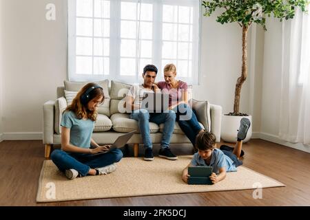 Giovane famiglia caucasica con padre, madre, figlia e figlio che usa un computer portatile, un tablet digitale e un telefono cellulare per navigare nei social media in salotto a casa Foto Stock