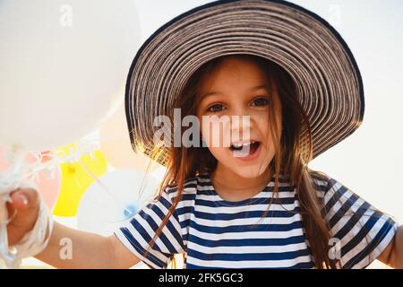 Primo piano divertente ritratto di ridere carino piccola ragazza tenendo palloncini colorati in cappello a righe e camicia guardando la macchina fotografica Foto Stock