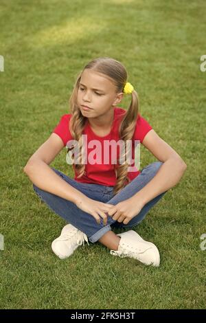 Perché così serio. Il bambino si siede su erba verde. La bambina indossa capelli lunghi. Piccolo bambino in stile estivo. Bellezza aspetto del bambino piccolo. Bambini Foto Stock