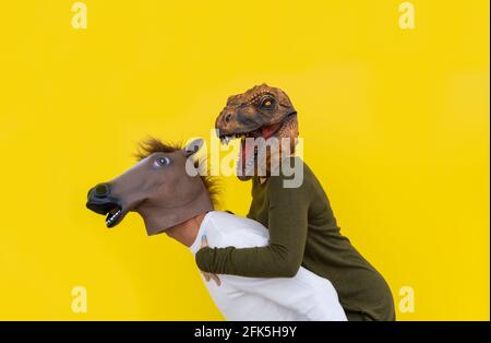 Felice coppia ritratto di uomo con testa di cavallo piggibacking sulle spalle donna indossando maschera di dinosauro testa isolato su sfondo giallo. Foto Stock