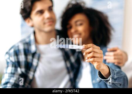 Gioiosa confusa giovane coppia sposata multirazziale in abiti casual godendo della buona notizia, preparandosi a diventare genitori, felice ragazza afroamericana tiene un test di gravidanza positivo Foto Stock