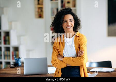 Ritratto di una bella ragazza afroamericana felice con capelli ricci in abiti eleganti, freelance, studente o manager, in piedi vicino alla scrivania, con le braccia incrociate, guarda al lato, sorridente piacevole Foto Stock