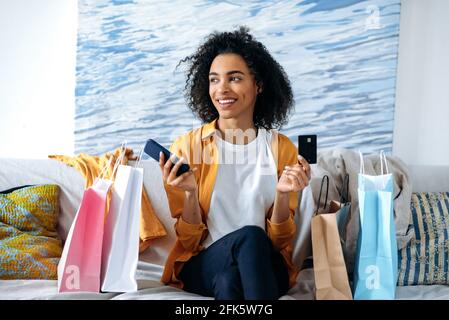 Felice bella donna afroamericana con capelli ricci, seduta sul divano, felice con nuovi acquisti, utilizza smartphone e carta di credito per i pagamenti online, molte borse di carta accanto ad essa, guarda al lato, sorride Foto Stock