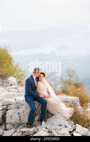 La sposa e lo sposo sono seduti abbracciando una roccia sul Monte Lovcen, dietro di loro si trova la Baia di Cattaro Foto Stock