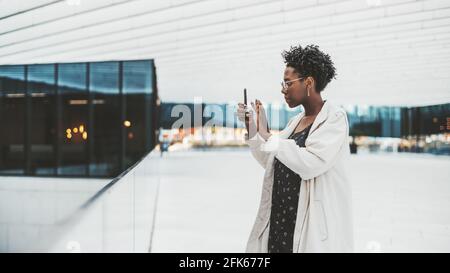 Vista laterale di una giovane bella femmina nera in occhiali, trench bianco, e con l'arte delle unghie, scattando foto in una galleria di un business moderno di Foto Stock