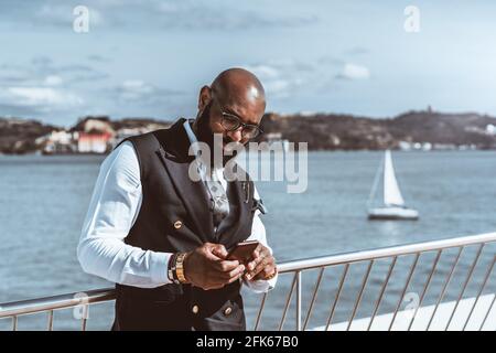 Ritratto di un dapper bearded uomo nero imprenditore calvo in un elegante gilet come parte del suo costume alla moda, utilizzando il suo cellulare in piedi n Foto Stock