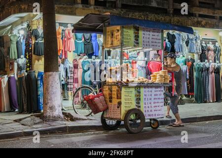 Carrello alimentare vietnamita che vende torte e panini parcheggiati di fronte al negozio di abbigliamento al crepuscolo, Hoi An Foto Stock