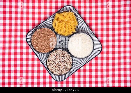 Cibo vegetale. Un piatto decorativo con quattro ciotole con grano saraceno, tagliatelle, riso e quinoa su una tovaglia a scacchi rossi. Mangiare sano. Vista dall'alto wi Foto Stock