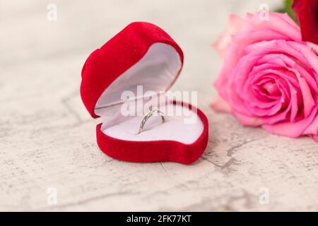 anello di nozze con diamante in una scatola a forma di cuore rosso e un bouquet di rose su uno sfondo bianco testurizzato. Concetto di impegno, dono, anniversario, Foto Stock