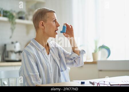 Vista laterale della donna di mezza età in abbigliamento casual con inalatore mentre ha attacco di asma, seduto al tavolo in cucina a casa, fuoco selettivo sulla donna anziana. Condizioni mediche e concetto di assistenza sanitaria Foto Stock