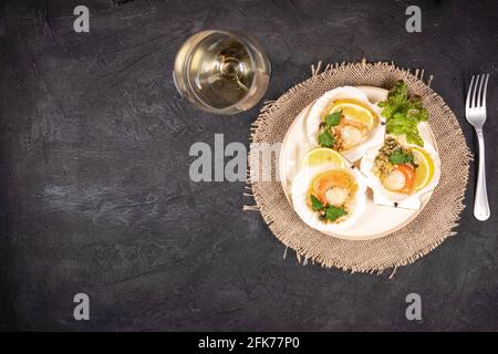 Capesante al forno con caviale in un piatto e un bicchiere di vino bianco. Capesante con limone su fondo nero con tovagliolo e forchetta. Messa a fuoco morbida Foto Stock