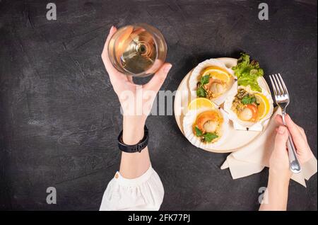 Capesante al forno con caviale e un bicchiere di vino su fondo nero. Le mani femminili tengono un bicchiere di vino bianco e una forchetta accanto alle capesante cotte con c Foto Stock