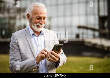 Felice uomo d'affari senior è rilassante dopo il lavoro di fronte alla costruzione dell'azienda. Sta utilizzando lo smartphone per la messaggistica. Foto Stock