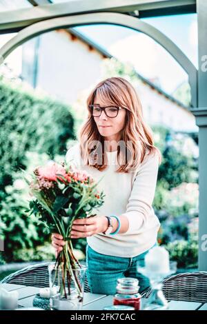 Foto di bella donna di mezza età in piedi nel balcone a casa mentre si dispone fiori sul tavolo. Foto Stock