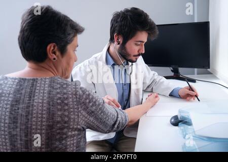 Giovane medico che ascolta la donna matura paziente cuore con uno stetoscopio e scrivere. Esame medico. Foto Stock