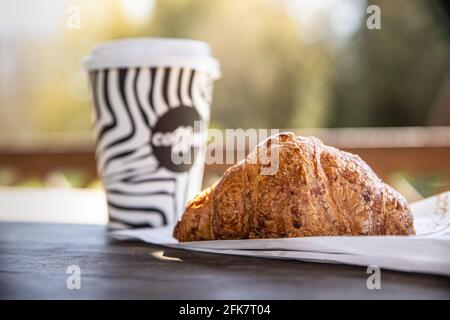 Tazzina monouso da asporto con caffè e croissant fresco su panca di legno nel giardino del mattino. Caffè in movimento. Natura sfondo. Messa a fuoco selettiva. Foto Stock