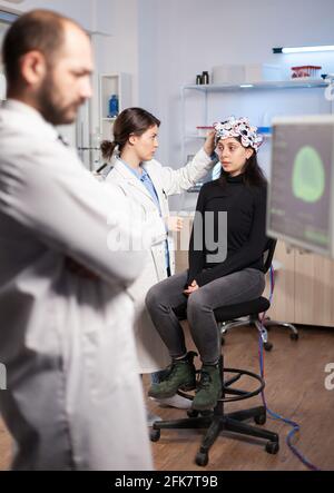 Neuroscience scientist che regola le cuffie con sensori su pazienti womna in laboratorio moderno durante la scansione cerebrale. Analisi del sistema nervoso, attrezzature cliniche. Scansione del cervello del medico. Foto Stock