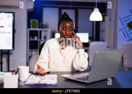 Donna d'affari africana che discute con il partner a tarda notte dall'ufficio. Occupato freelance focalizzato che fa straordinari per la scrittura di lavoro di lettura, la ricerca, rispettando la scadenza del progetto finanziario, overworking. Foto Stock