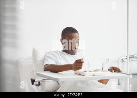 Ritratto dell'uomo afroamericano che mangia cibo in ospedale mentre giace su letto bianco, copia spazio Foto Stock
