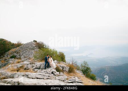 La sposa e lo sposo sono seduti abbracciando una roccia sul Monte Lovcen, dietro di loro si trova la Baia di Cattaro Foto Stock