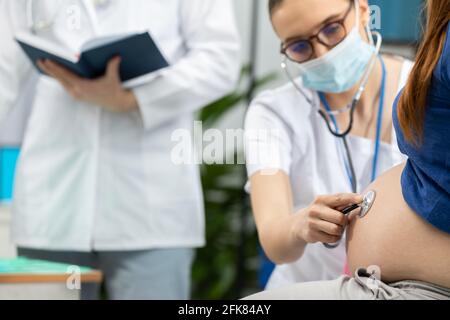 Un medico di signora che indossa una maschera protettiva esamina l'addome con uno stetoscopio di una paziente incinta. Durante la pandemia. Foto Stock