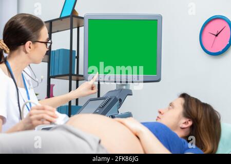 Un medico mostra a una paziente incinta il suo bambino non ancora nato su uno schermo a ultrasuoni. Schermo verde. Ufficio di Ginecologia. Foto Stock