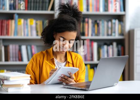 Gioiosa intelligente concentrata afroamericana studentessa, prendendo appunti in un notebook, ascoltando le lezioni online, guadagnando conoscenza, mentre si siede al tavolo nella biblioteca universitaria, sorridendo Foto Stock