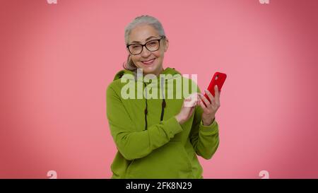 Anziana donna Granny utilizzare il telefono cellulare di navigazione online dire wow sì, grande vincere notizie facendo gesto vincitore Foto Stock