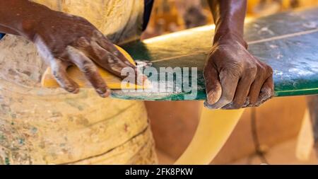 Uno Shaper da surf di un artigiano africano che lavora in un'officina di riparazione Con carta vetrata Foto Stock