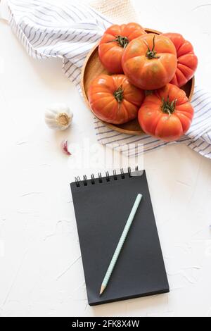 Mazzo di pomodori in un vassoio di legno su un tovagliolo rustico su sfondo bianco. Un blocco note a spirale nera aperto è pronto per essere scritto con una ricetta. Foto Stock