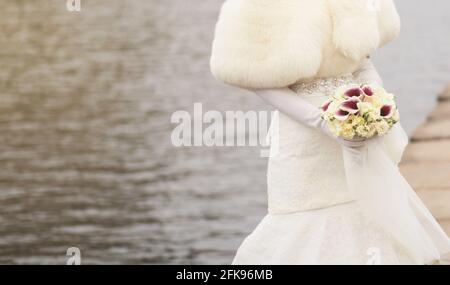 corride in un cappotto di pelliccia con un bouquet di nozze sullo sfondo del fiume. copia spazio Foto Stock