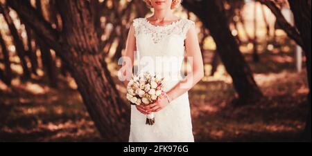 sposa in un abito bianco che tiene un bouquet di fiori sullo sfondo di erba verde Foto Stock