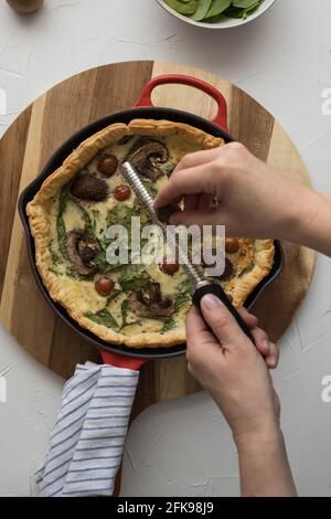 La donna grattugia una noce moscata su una quiche torrefatta a caldo all'interno di una padella metallica sopra un tagliere di legno. Foto Stock